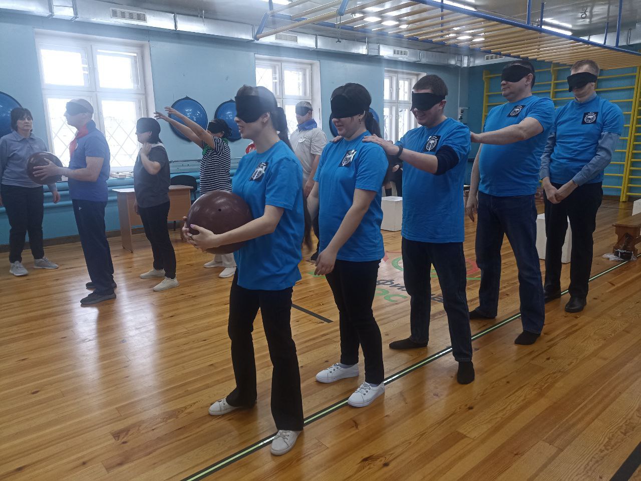 Тифлокомментарий к фотографии: В спортивном зале Бийского филиала ЦРС ВОС команды выстроены в  две колонны. В руках впереди стоящих игроков большой мяч. Команды готовы к выполнению задания.