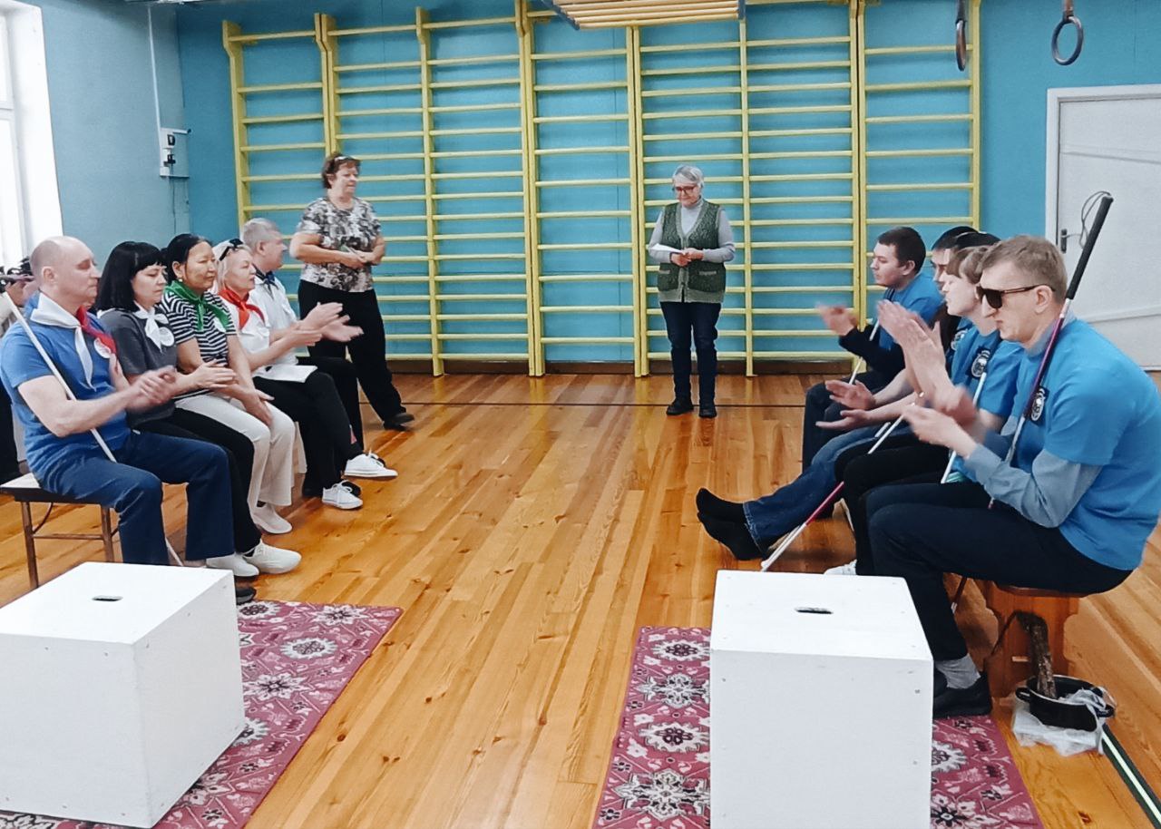 Тифлокомментарий к фотографии 1: В спортивном зале Бийского филиала ЦРС ВОС напротив друг друга сидят две команды участников конкурса. В руках сидящих людей белые трости. Команда справа в одинаковых футболках ярко-синего цвета, команда слева в разноцветных шейных косынках-галстуках. На заднем плане расположена шведская стенка, перед которой находится ведущий мероприятия с листом бумаги в руках.