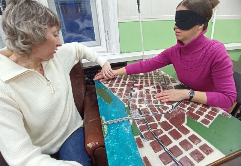 Тифлокомментарий: на фотографии две женщины, они сидят за столом напротив друг друга. На столе перед ними расположен рельефный план местности. На глазах одной из женщин надета черная повязка – она активный участник мастер-класса. Вторая женщина – преподаватель, она своей левой рукой направляет правую кисть обучающегося по элементам плана.