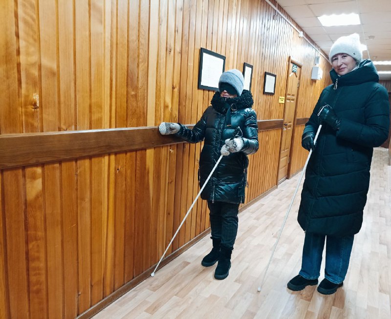 Тифлокомментарий: Две женщины в верхней зимней одежде стоят в коридоре лицом к камере. На глазах одной из них черная маска и белая трость в руках – она активный участник мастер-класса, вторая женщина с белой тростью в руках – преподаватель, она смотрит в камеру, улыбается. 